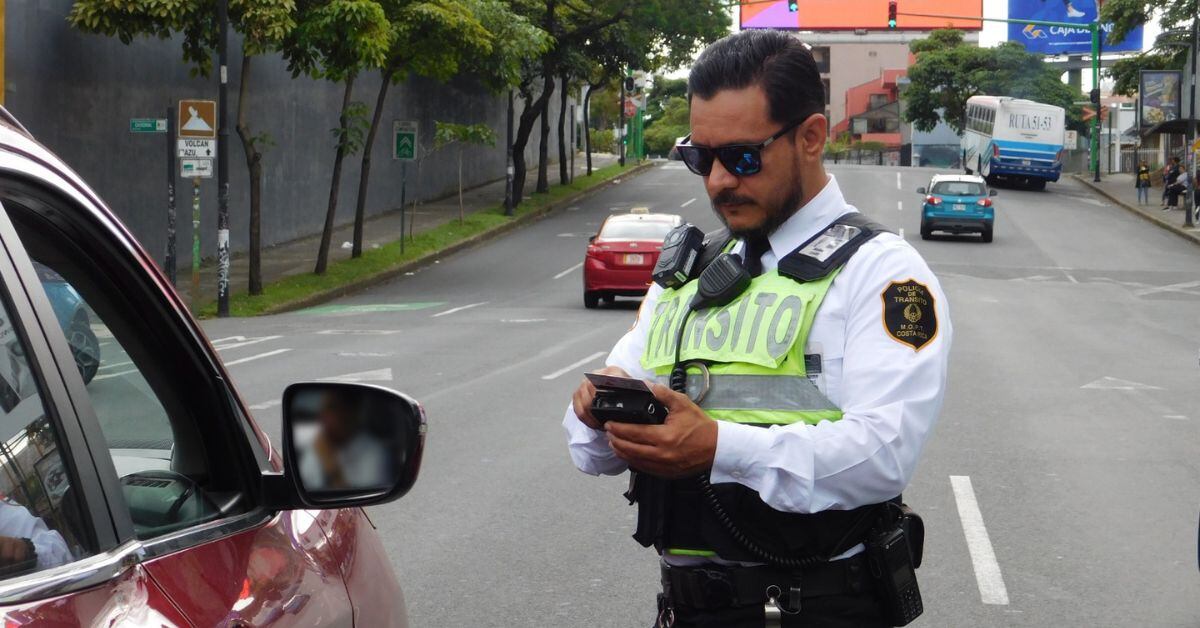 Un oficial de Tránsito con chaleco reflectante revisa un dispositivo handheld frente a un vehículo detenido en una calle de Costa Rica.