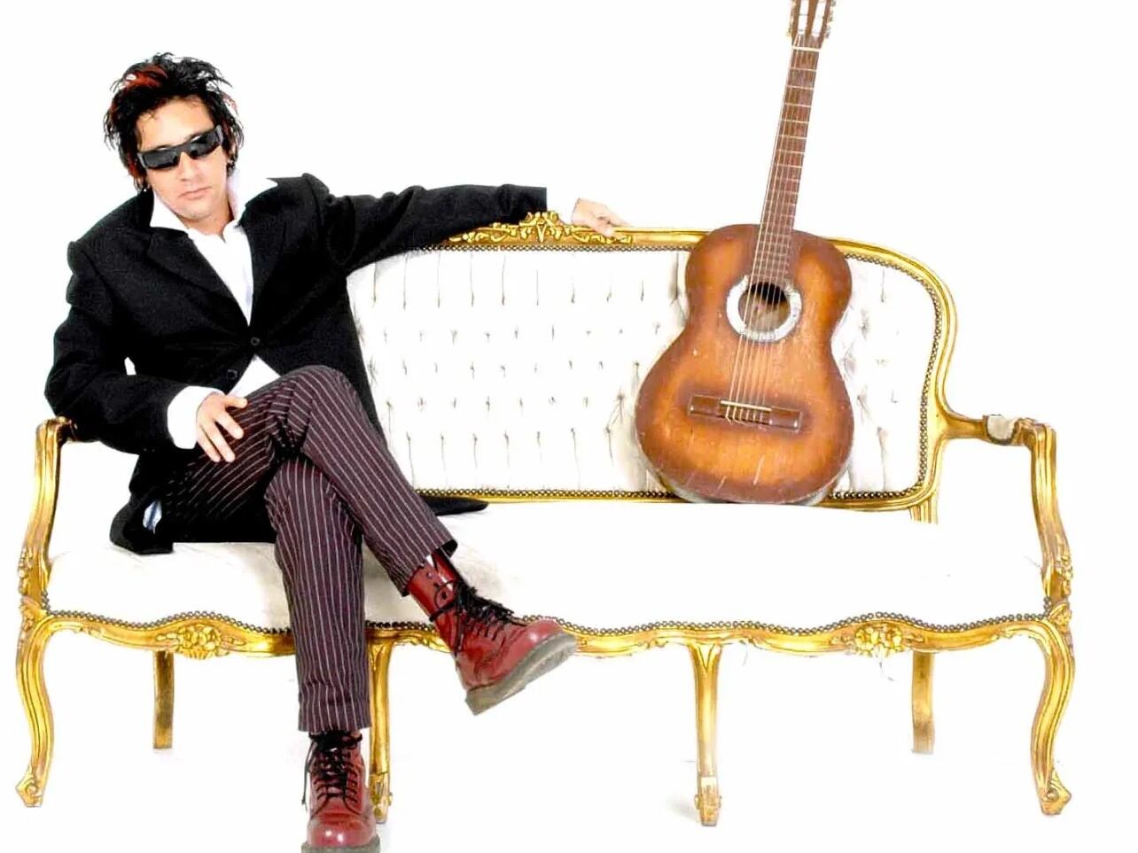 Luis Lauretto sentado en un sillón blanco con la pierna cruzada y una guitarra al lado.