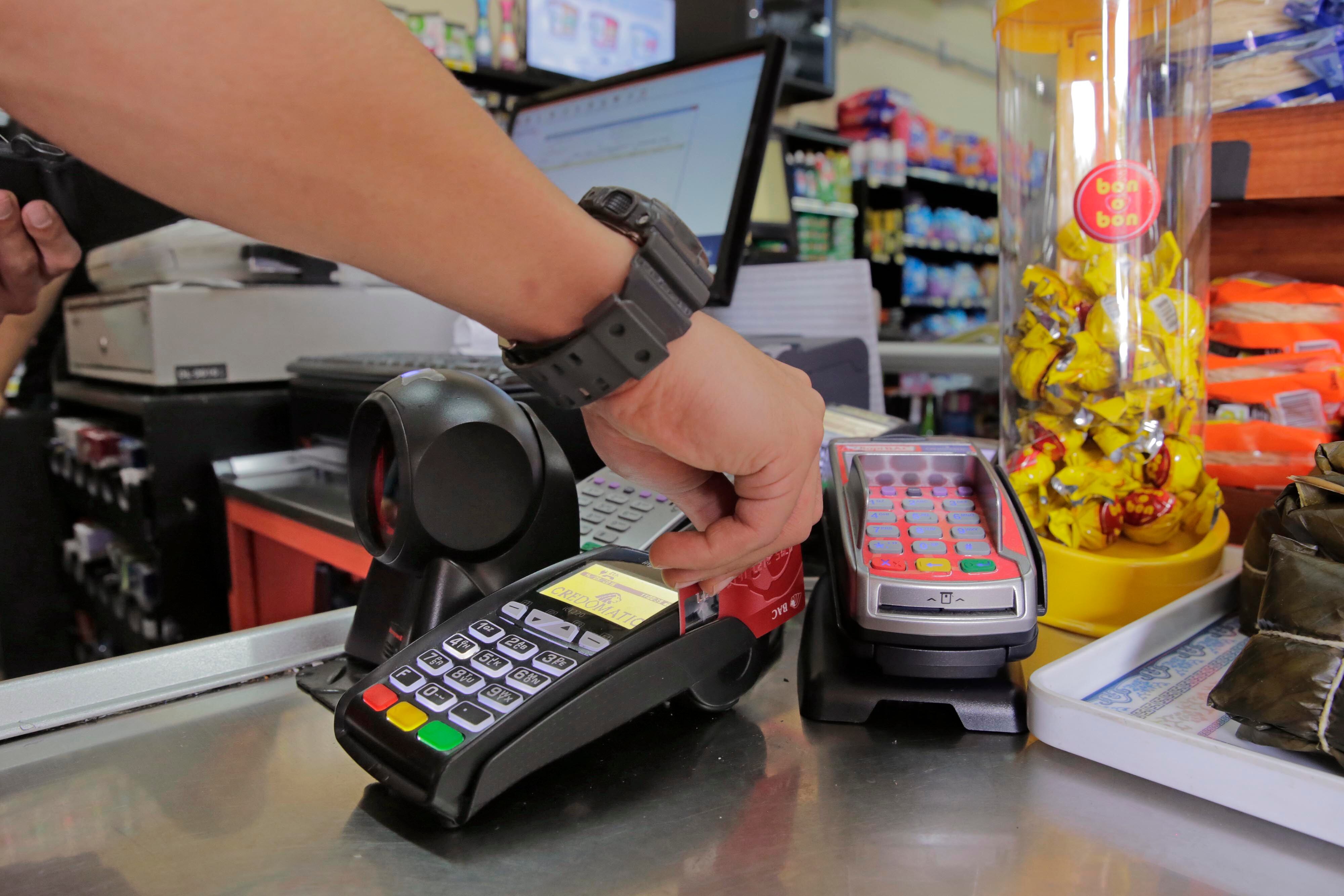 Mano de una persona insertando una tarjeta en un datáfono en un comercio, con otros dispositivos y productos visibles en el fondo.