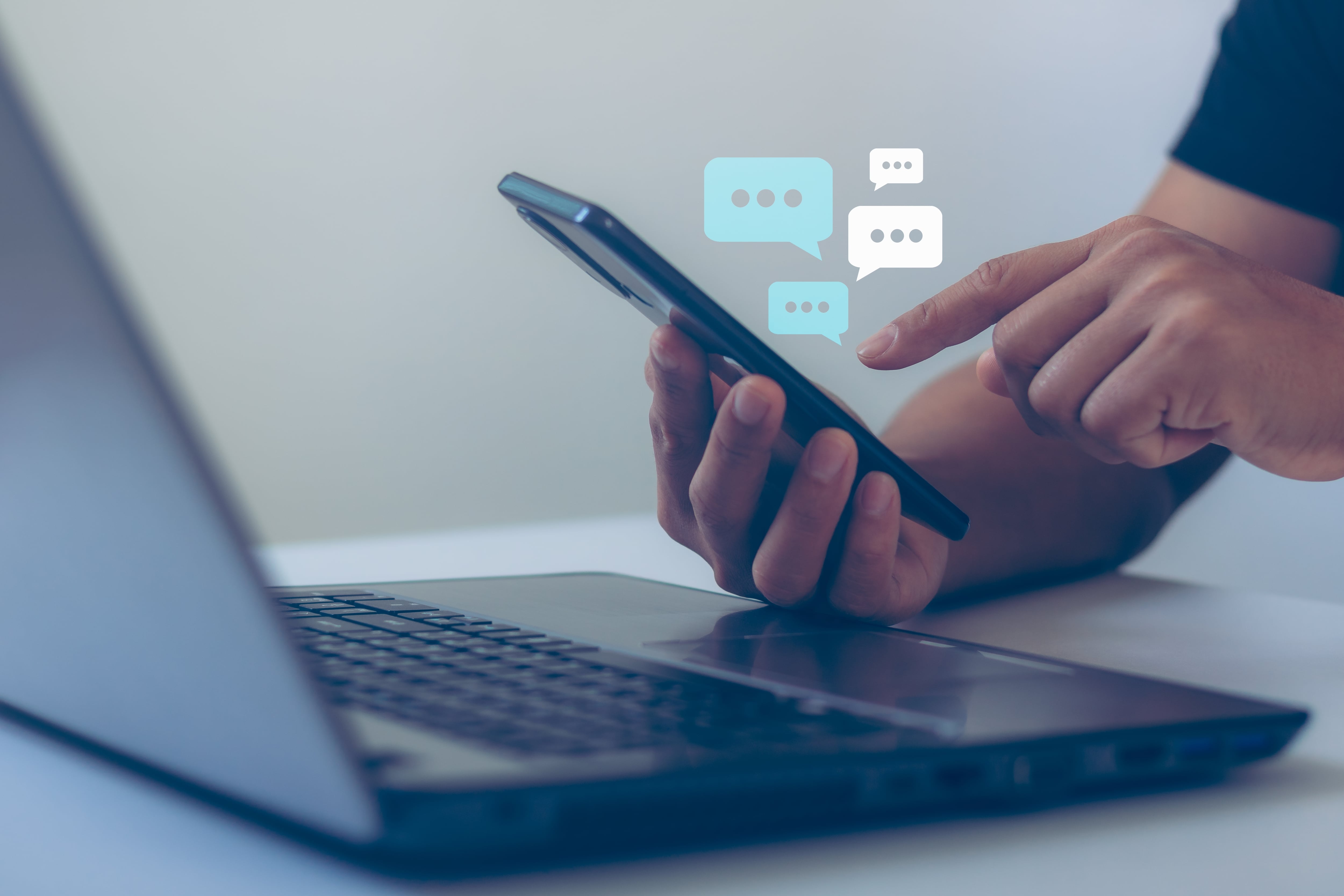 Manos de hombre tecleando en su celular y una laptop, computadora portátil sobre la mesa, mensajería instantánea