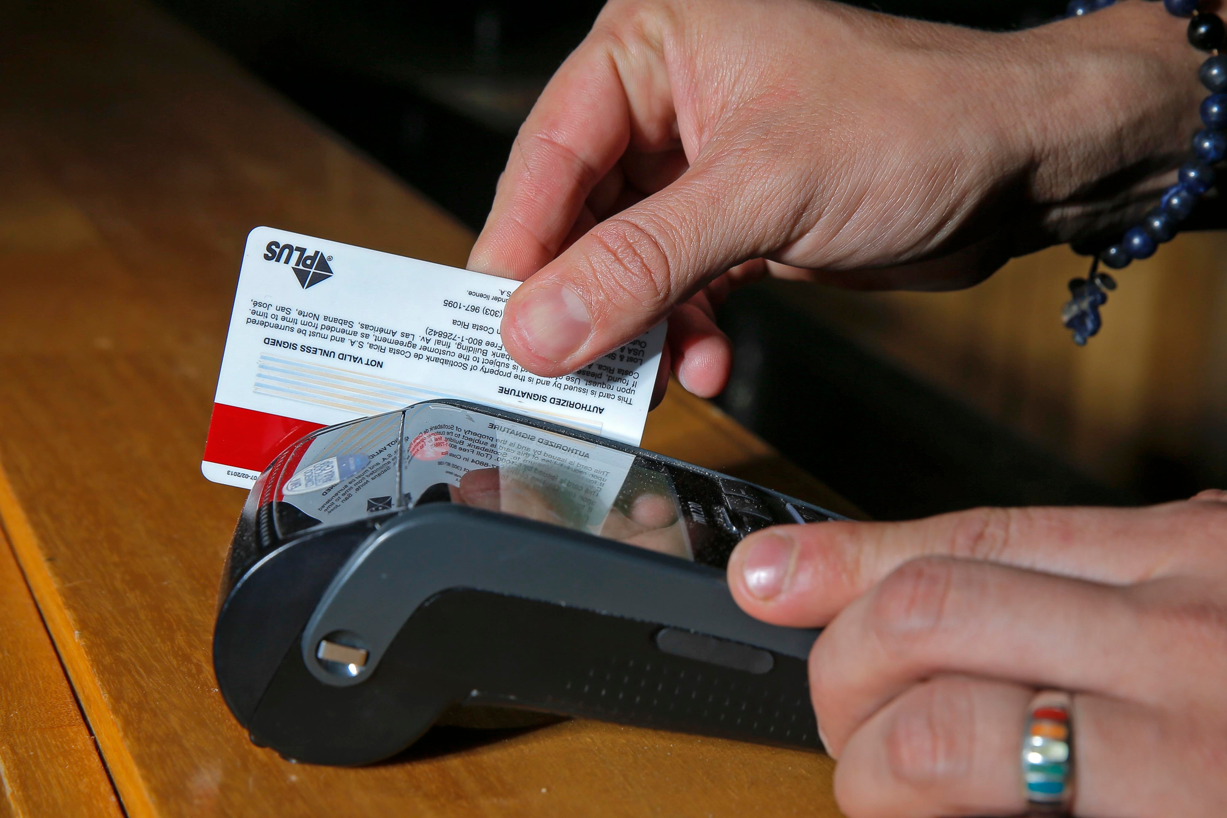 Mano con anillos, detalle de los dedos sosteniendo una tarjeta de plastico, que pasa por un aparato negro conocido como datáfono.