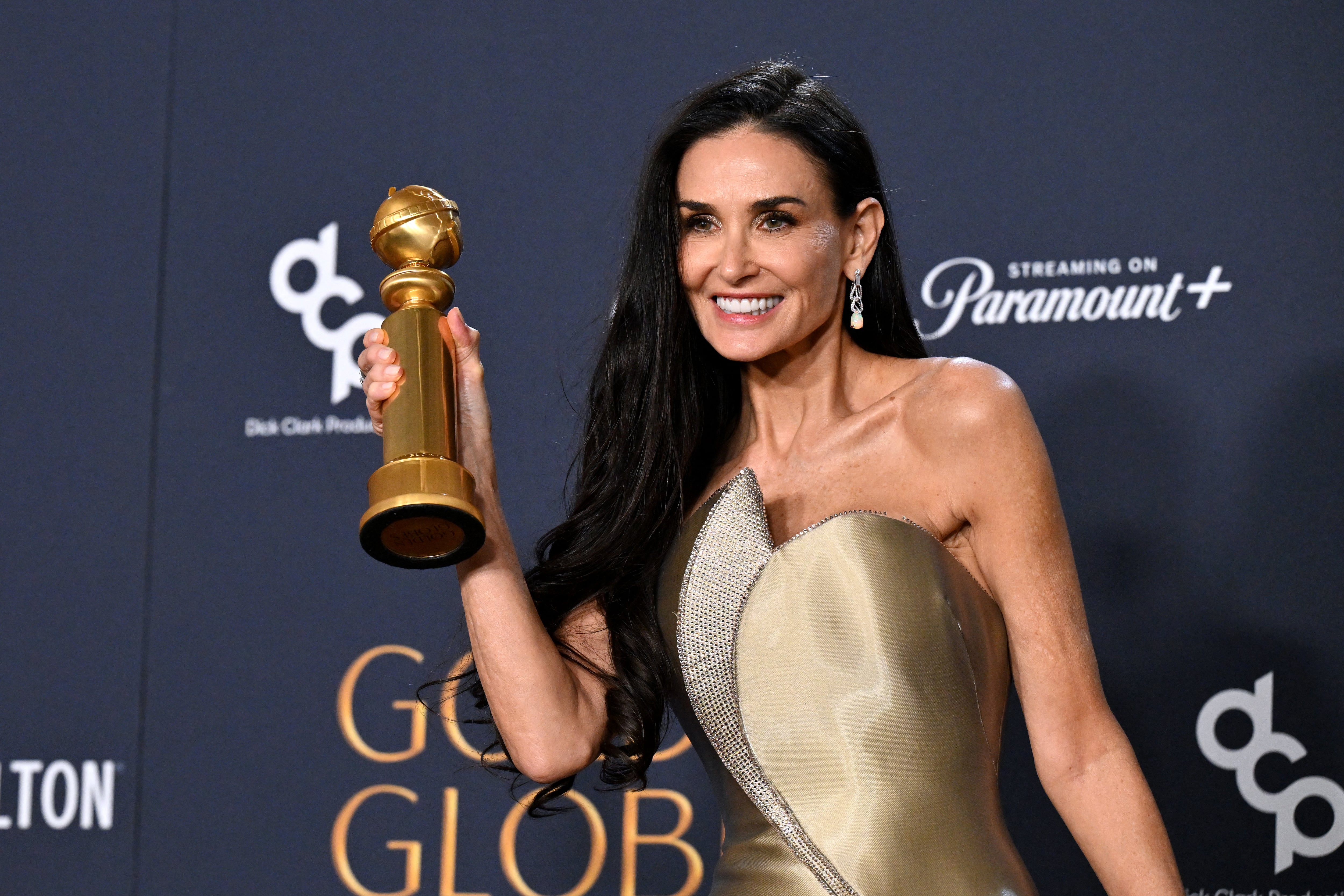Imágenes de la actriz Demi Moore sosteniendo en su mano derecha un premio Globo de Oro. Ella sonríe, su cabello negro largo va suelto y luce un vestido largo en tono dorado.