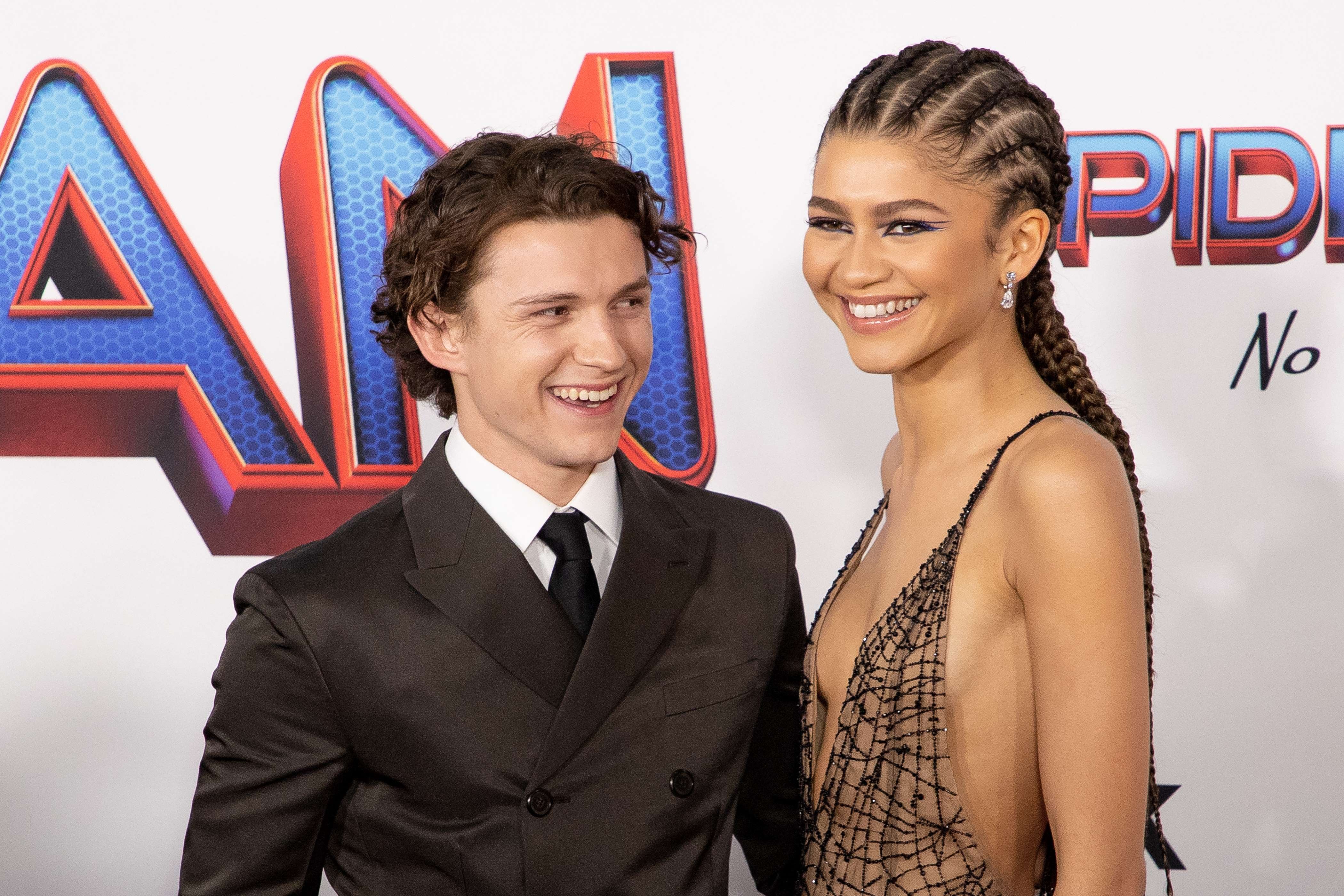 Foto en la que se ven juntos los actorez Tom Holland y Zendaya sonriendo. Él usa un traje de tono oscuro con corbata, ella un vestido con amplio escote y trenzas en su cabello.
